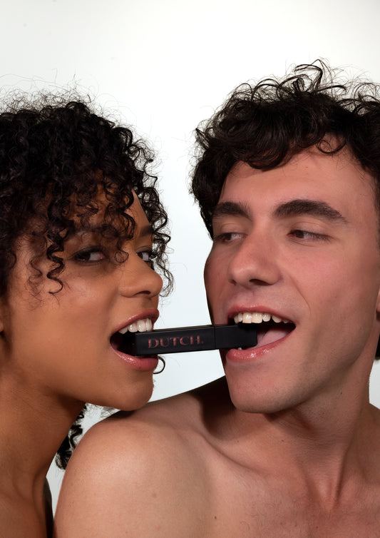 Woman and man holding a lipgloss tube in their mouths. It is a matte black tube with the DUTCH. logo transparant in it.