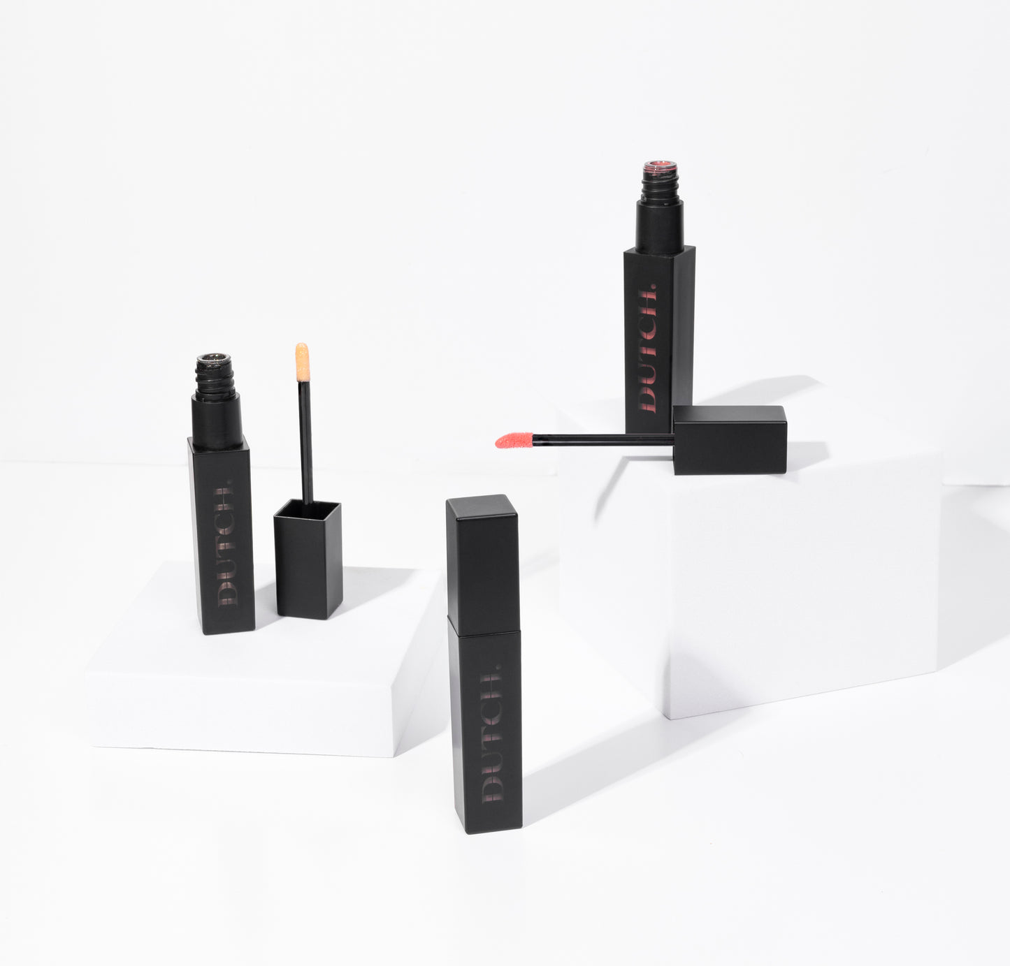Three tubes of lipgloss in different colors. The tubes are matte black with DUTCH. transparant shown in it. De lipgloss colors are called Hope (transparant), High (bronze) and Hype (coral).